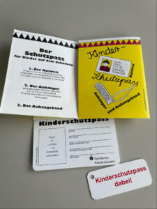 „Aufgeklappter Kinderschutzpass mit Platz für Lichtbild und persönlichen Angaben wie Name, Geburtsdatum und Adresse. Der Ausweis aus Kunststoff ist beidseitig beschriftbar und versiegelbar. Darunter liegt ein Anhänger mit der Aufschrift ‚Kinderschutzpass dabei!‘ und einem robusten Anhängeband.